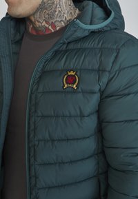Mörkgrön vadderad jacka med en zipprad huva, med horisontell sömnad och en broderad logotyp i röd och guld på bröstet.