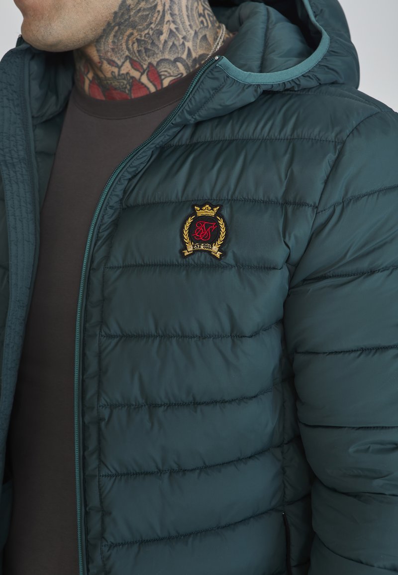 Giacca imbottita verde scuro con cappuccio con zip, con cuciture orizzontali e un logo ricamato in rosso e oro sul petto.