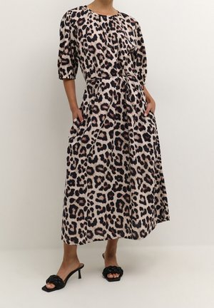 Femme portant une robe midi à imprimé léopard avec des manches bouffantes courtes, une ceinture à la taille, des poches latérales et des sandales noires tressées à talons.