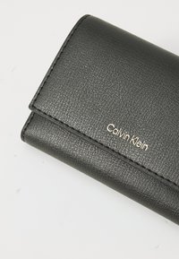 Svart teksturert skinnlommebok fra Calvin Klein som er brettet igjen, med synlig merkevarelogo på en hvit bakgrunn.