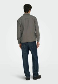 Camisa gris de botones con mangas largas, con costura en la parte posterior y puños, combinada con pantalones de mezclilla azul oscuro y zapatos negros.
