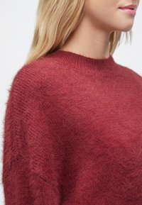 Κρεμ κόκκινο πλεκτό πουλόβερ με απαλό, χνουδωτό υφή, στρογγυλή λαιμόκοψη και ελαφρώς φαρδιά εφαρμογή. Το ύφασμα διαθέτει διακριτικό μοτίβο και ριμπ μανσέτες.