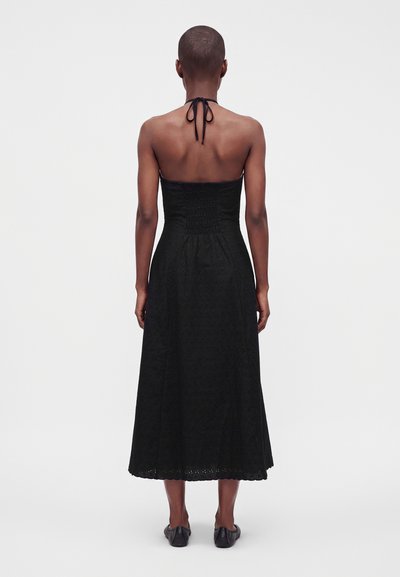 Γυναίκα με κοντά μαλλιά, ντυμένη με μαύρο strapless halter midi φόρεμα και μαύρα παπούτσια, στέκεται με την πλάτη προς ένα απλό λευκό φόντο.