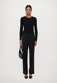 Haut noir à manches longues associé à un pantalon noir à rayures, tenant un sac à main noir. Les chaussures ont des bouts pointus. Design simple et ajusté.