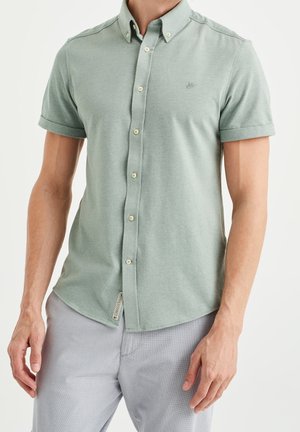 Uomo che indossa una camicia verde chiaro a maniche corte con bottoni e polsini arrotolati e pantaloni grigio chiaro su sfondo semplice.