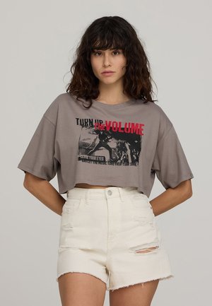 T-shirt grigia corta con maniche corte, caratterizzata da un audace testo grafico in nero e rosso. Abbinata a pantaloni corti bianchi a vita alta e strappati.