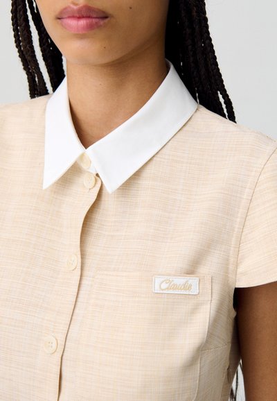 Chemise à manches courtes jaune pâle avec un col blanc, boutonnage sur le devant, et une petite poche plaquée avec une étiquette brodée. Tissu texturé.