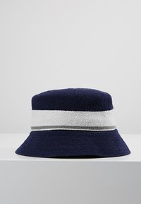 Chapeau seau en terry bleu marine avec un large bande rayée blanche et grise. Forme ronde et douce avec un bord flexible. Matériau texturé et pelucheux.