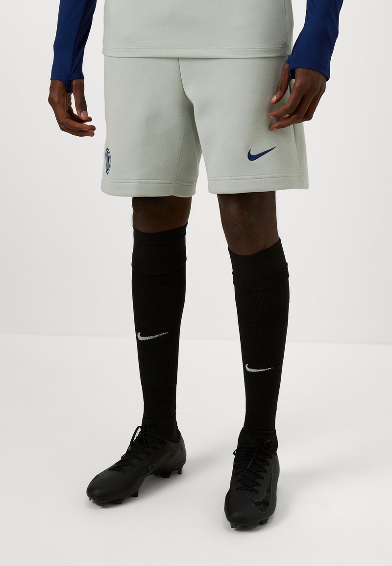 Lys grå Nike shorts med et blåt logo, kombineret med sorte knæhøje sokker med et hvidt logo og sorte fodboldstøvler.