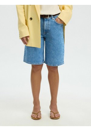 Osoba nosząca niebieskie dżinsowe spodenki za kolano, żółtą marynarkę i beżowe sandały na obcasie, stojąca z jedną ręką w kieszeni.