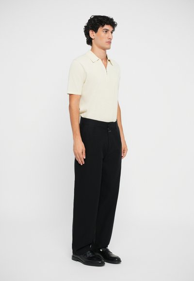 Polo corto de color beige claro con cremallera, combinado con pantalones negros. El conjunto tiene un corte relajado y una textura suave. Unos zapatos negros completan el look.