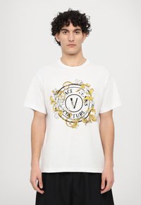 Balti medvilnės marškinėliai su apvalia gėlių ornamentika, juodos ir geltonos spalvų akcentais, su užrašu "Versace Jeans Couture."