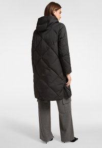 Vrouw in een lange zwarte gewatteerde jas met een capuchon, gecombineerd met grijze wijde broekspijpen en zwarte hakken, staand met haar zij naar de camera.