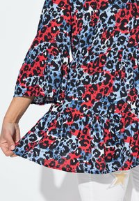 Blusa con stampa leopardata rossa, blu e nera, con maniche a campana e orlo a volant a più strati. Tessuto leggero e testurizzato. Primo piano di una mano che tiene l'orlo.