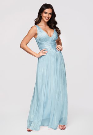 Femme aux longs cheveux foncés portant une robe plissée bleu clair sans manches, debout les mains sur les hanches et souriant sur un fond uni.