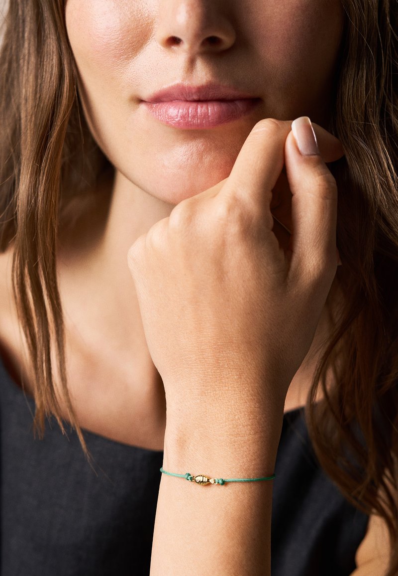 Gros plan d'une femme touchant son menton avec la main, portant un bracelet fin vert avec des perles dorées au poignet.