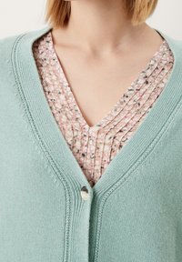 Světle zelený cardigan s výstřihem do V a jedním knoflíkem, navlečený přes skládací blůzu s květinovým vzorem v jemných pastelových barvách.