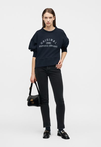 Γυναίκα στέκεται φορώντας μαύρο φούτερ με την επιγραφή "Original 1981 Emporio Armani", μαύρο στενό τζιν, μαύρες μοκασίνες, και κρατώντας μαύρη τσάντα, με φόντο λευκό.