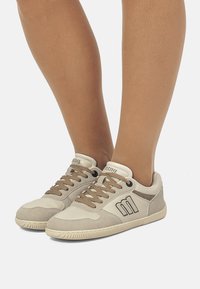 Beige Sneakers mit einem weichen Obermaterial aus Wildleder und Leder, das braune Schnürsenkel, eine strukturierte Sohle und ein Logodetail an der Seite aufweist.