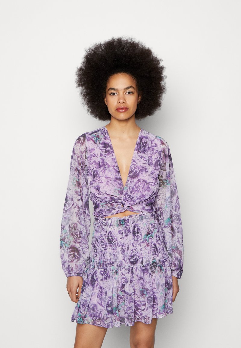 Vestido de estampa floral roxa com mangas longas e transparentes, decote em V profundo e detalhe frontal com nó. A saia tem um design de camadas com babados.