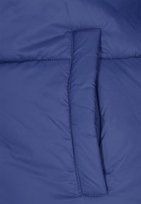 Gros plan d'une veste matelassée bleu marine avec une texture en tissu quilté et un détail de poche à rabat. Le matériau semble lisse avec un léger éclat.