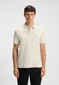 Beige polo shirt met een korte kraag, voorzien van een hexagonaal logo in lichtbruin op de borst. Zacht, gladde textuur, standaard fit.