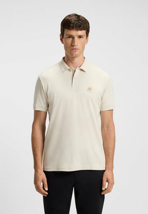 Beige Poloshirt mit kurzem Kragen, versehen mit einem hexagonalen Logo in Hellbraun auf der Brust. Weiche, glatte Textur, normale Passform.
