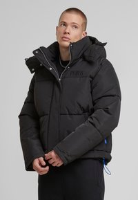 Černá pouzdrová bunda s vysokým límcem, zipem vpředu a detaily loga na hrudi. Má elastické manžety a stažený spodní lem.