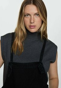 Grijze mouwloze coltrui gecombineerd met zwarte denim salopettes. Zichtbare schouderbanden en een gladde textuur; neutraal kleurenpalet.