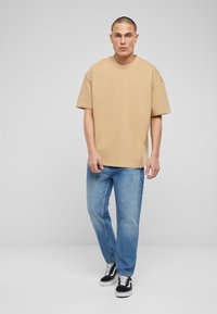 Camiseta de algodón beige con mangas cortas anchas, combinada con jeans de mezclilla azul claro. El calzado incluye zapatillas negras y blancas con un diseño de cordones.