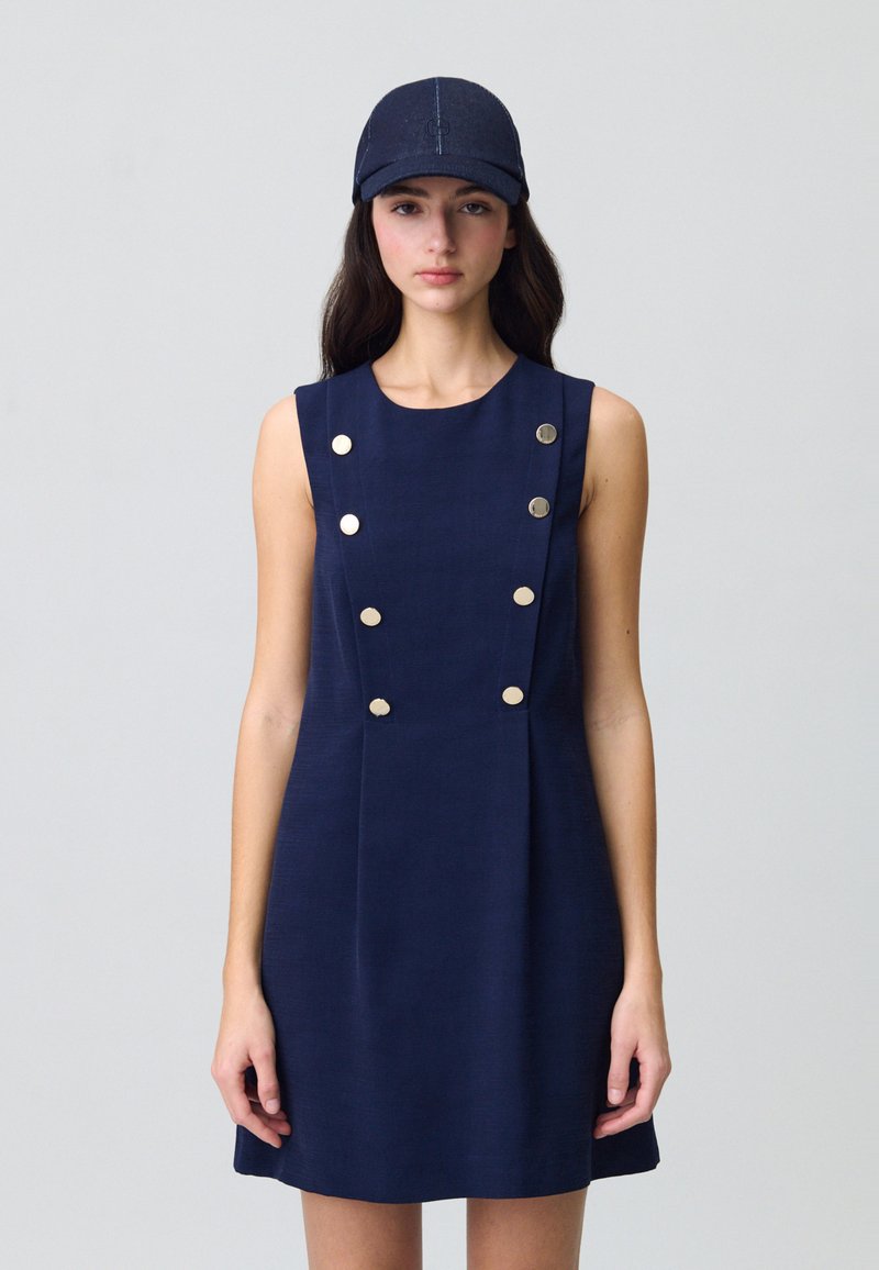 Vestido sin mangas de color azul marino con una textura suave y seis botones dorados en el frente. Combinado con una gorra a juego de color azul marino, también texturizada.
