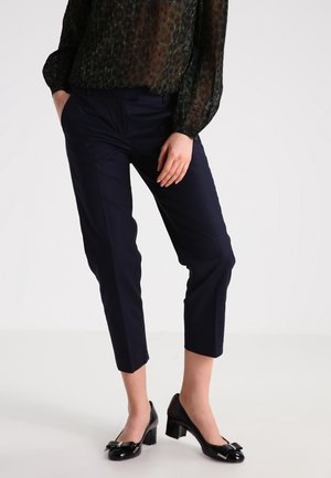 Kvinde iført sorte cropped bukser, sort gennemsigtig bluse med lange ærmer og sorte hæle med sløjfer, stående med den ene hånd i lommen.