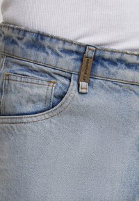 Šviesiai mėlyni džinsai su juosmeniu, priekinė kišenė ir matoma metalinė prekės ženklo etiketė ant diržo kilpos, dėvimi su balta šonkaine marškinėliais.