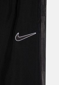 Melnas sporta bikses ar mīkstu tekstūru, ar baltu Nike logo uz kreisās augšstilba un ar rakstu sānu detaļām papildu efektam.