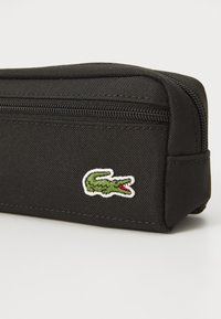 Trousse en toile noire avec fermeture éclair, ornée d'un logo de crocodile vert brodé en contour blanc sur le devant.