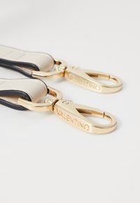 Leren band in crème met zwarte afwerking, voorzien van gouden metalen clips met de opdruk "VALENTINO", die een gladde textuur en strakke vormgeving tonen.