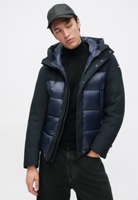 Doudoune navy avec capuche, composée d'un mélange de matériaux brillants et mats, fermeture éclair et boutons-pression. portée par-dessus un col roulé noir.
