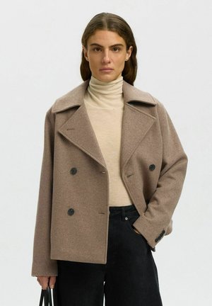 Vrouw draagt een beige dubbelgeknoopt wollen jas over een crèmekleurige coltrui, zwarte broek, staat met één hand in de zak en houdt een zwarte tas vast.
