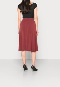 Jupe bordeaux, mi-longue, en tissu doux avec une taille élastique. Portée avec un haut noir et des chaussures à talons noirs.