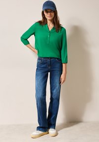 Blusa verde con cuello, de mangas tres cuartos, combinada con jeans acampanados azules. Zapatillas blancas con suelas beige y una gorra de béisbol azul marino.