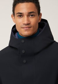 Giacca a vento navy con colletto alto, chiusura con zip e cinque bottoni neri per la chiusura. Il materiale sembra liscio e resistente all'acqua.