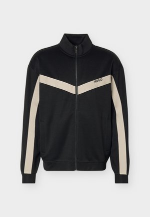 Veste noire zippée avec des bandes beige en forme de chevrons sur la poitrine et les manches, avec le logo "HUGO" sur la poitrine gauche.