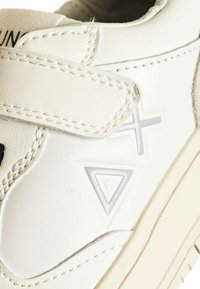Sneaker bianca in pelle con cinturino in velcro, caratterizzata da forme geometriche embossate e cuciture nere a contrasto. Texture liscia e design a profilo basso.