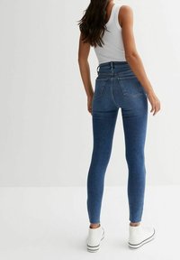 Mujer con jeans ajustados de cintura alta de color azul, camiseta sin mangas blanca y zapatillas altas blancas, de pie contra un fondo claro.