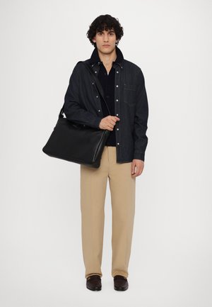 Geantă de umăr din piele neagră ținută de o persoană îmbrăcată cu o jachetă din denim, tricou negru și pantaloni bej deschis. Geanta are o textură netedă și un hardware minimal.