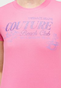 Κοντινό πλάνο σε ροζ μπλουζάκι με λαμπερό κείμενο "Versace Jeans Couture Beach Club" και μια μικρή εικόνα γοργόνας στη δεξιά πλευρά.