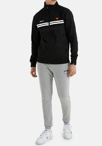 Zwarte zip-up jack met witte strepen, hoge kraag en geborduurd logo; gecombineerd met grijze joggingsbroek en witte sneakers.
