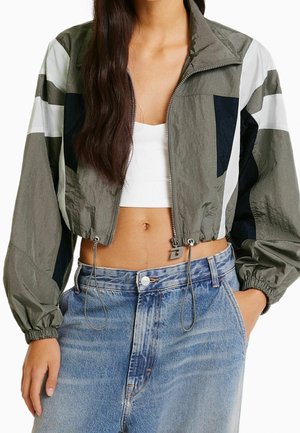 Mujer con chaqueta corta gris y negra con cremallera sobre un top corto blanco, combinada con jeans azules de talle alto, con una mano en el bolsillo.