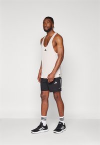 Top sin mangas de color rosa claro con un logo negro, combinado con pantalones cortos negros con rayas blancas. Lleva puestos zapatos deportivos negros y calcetas a rayas.