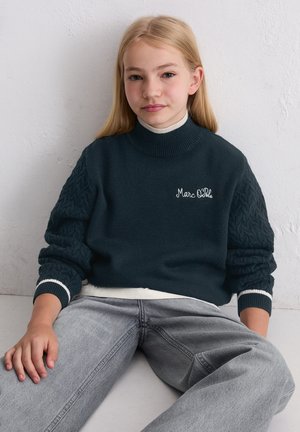 Girl with long blond hair sitting against a white wall, wearing a dark blue Marc O'Polo sweater and light gray jeans.